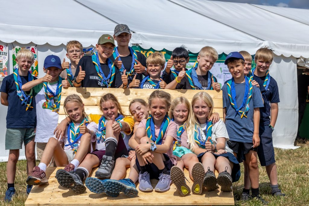 Glad flok juniorspejdere giver tommelfinger op foran et telt på Spejdernes Lejr 2026